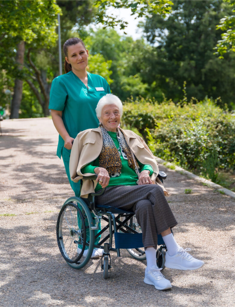 Eine ältere Dame im Rollstuhl wird von einer Pflegerin duch den Park gefahren.