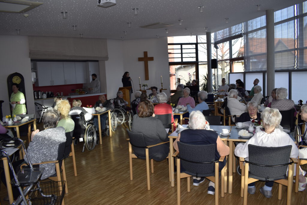 Bewohnerinnen und Bewohner sitzen an Tischen und hören die Begrüßungsworte