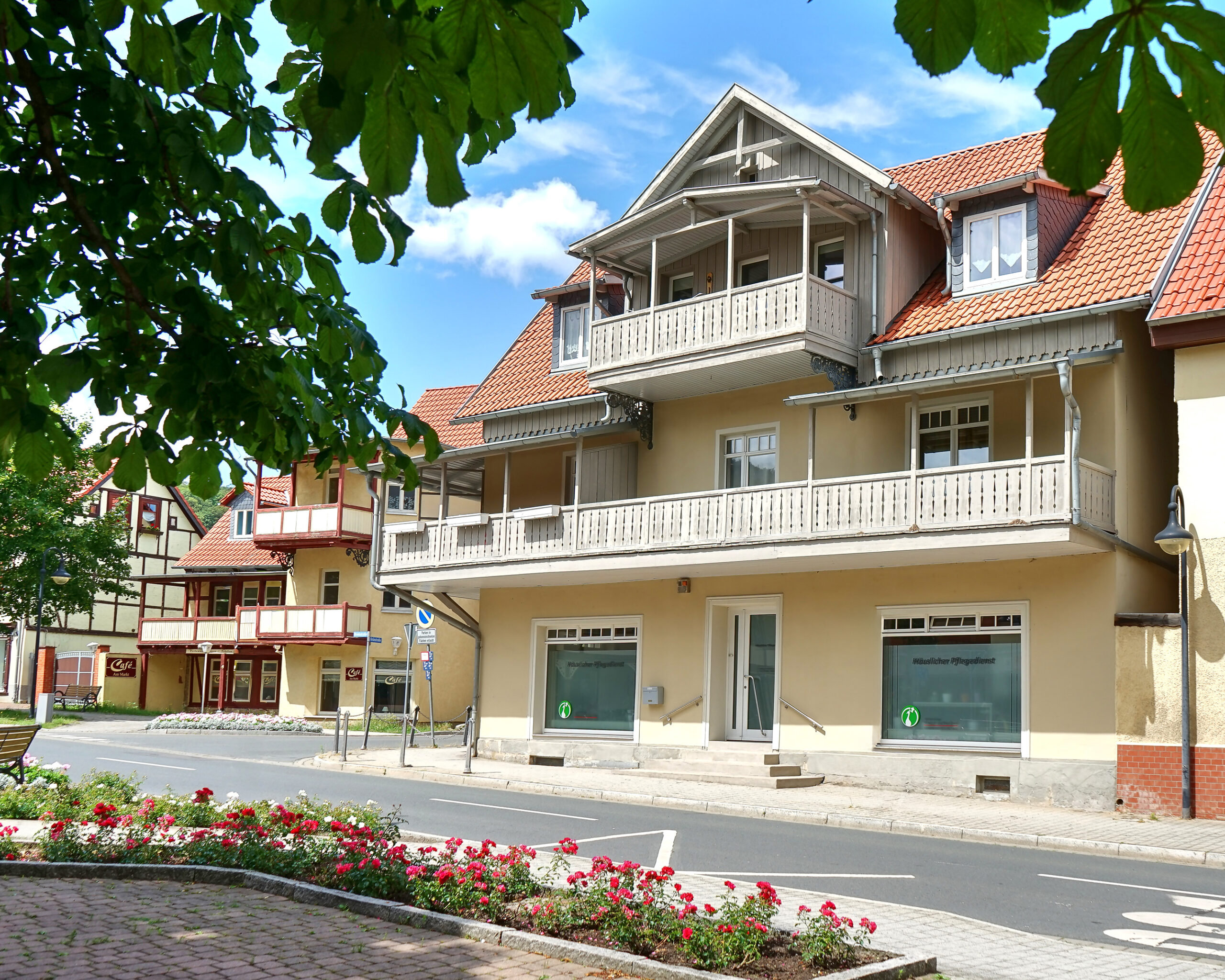 Gebäude mit Balkonen und rotem Ziegeldach an einer Straße; im Vordergrund ein Blumenbeet und Baumblätter.