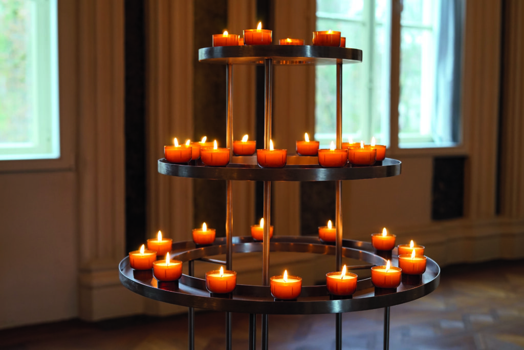 Mehrstöckiger Kerzenständer mit vielen brennenden Teelichtern in einem Raum vor Fenstern.