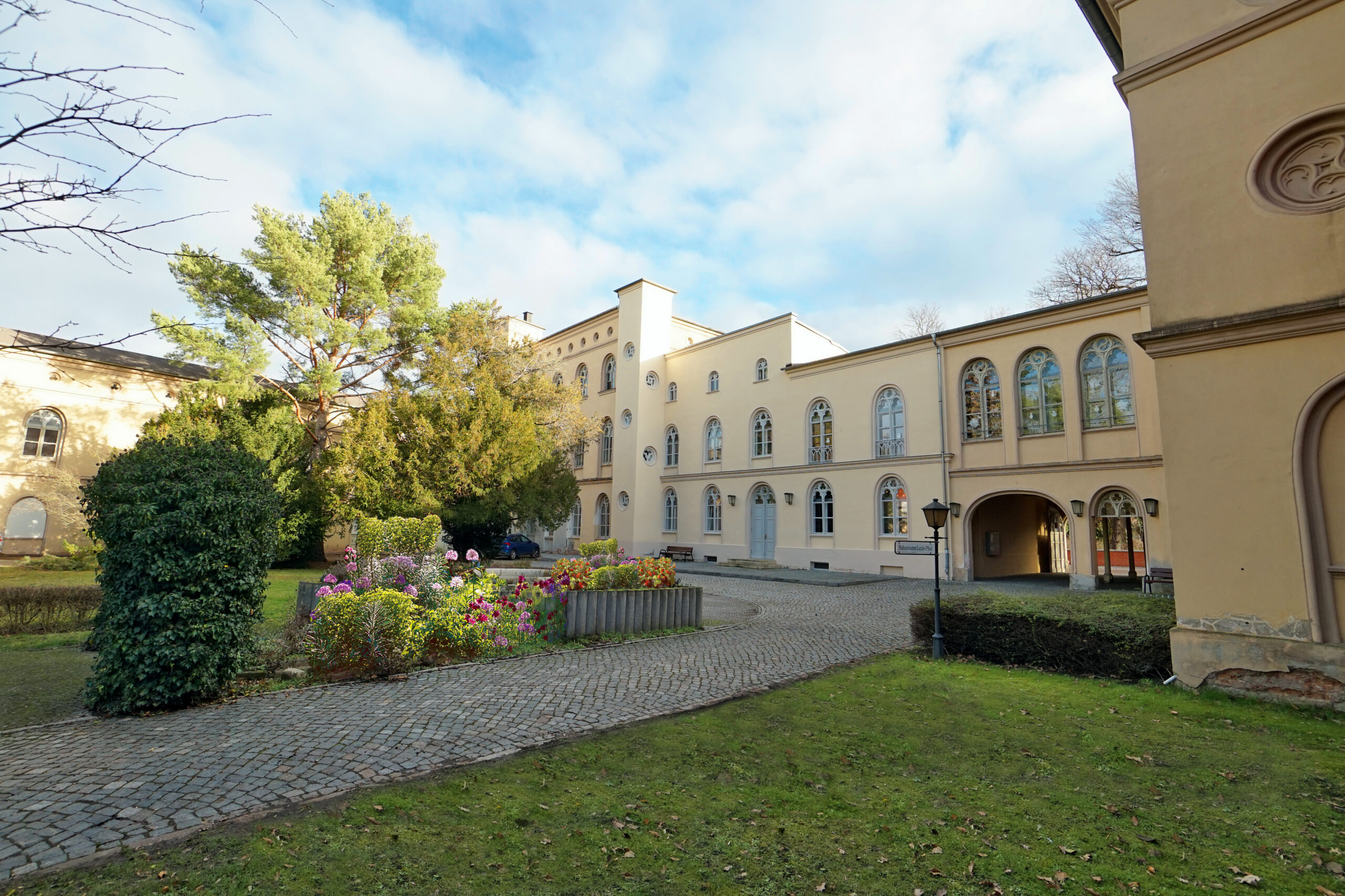 Heller Gebäudekomplex mit Rundbogenfenstern und Durchfahrt, davor ein gepflasterter Weg und ein Blumenbeet.